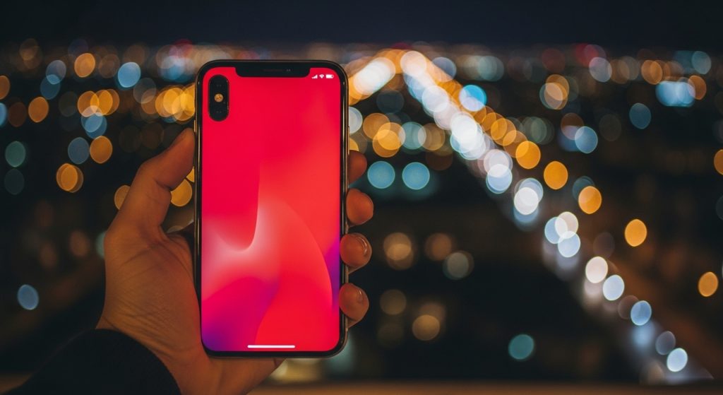 Aesthetic Wallpaper Background iPhone Red Bold & Stylish HD Designs 13 : A hand holding an iPhone with a striking red wallpaper, city lights blurred in the background.