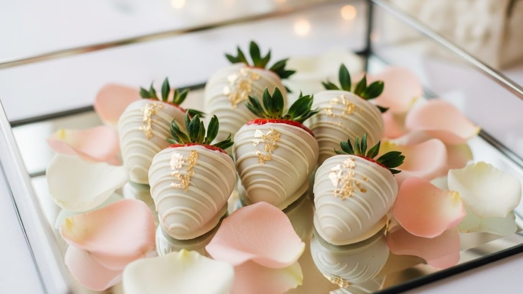 White chocolate-dipped strawberries touched with gold leaf, arranged on a mirrored tray with rose petals, glamorous bridal style