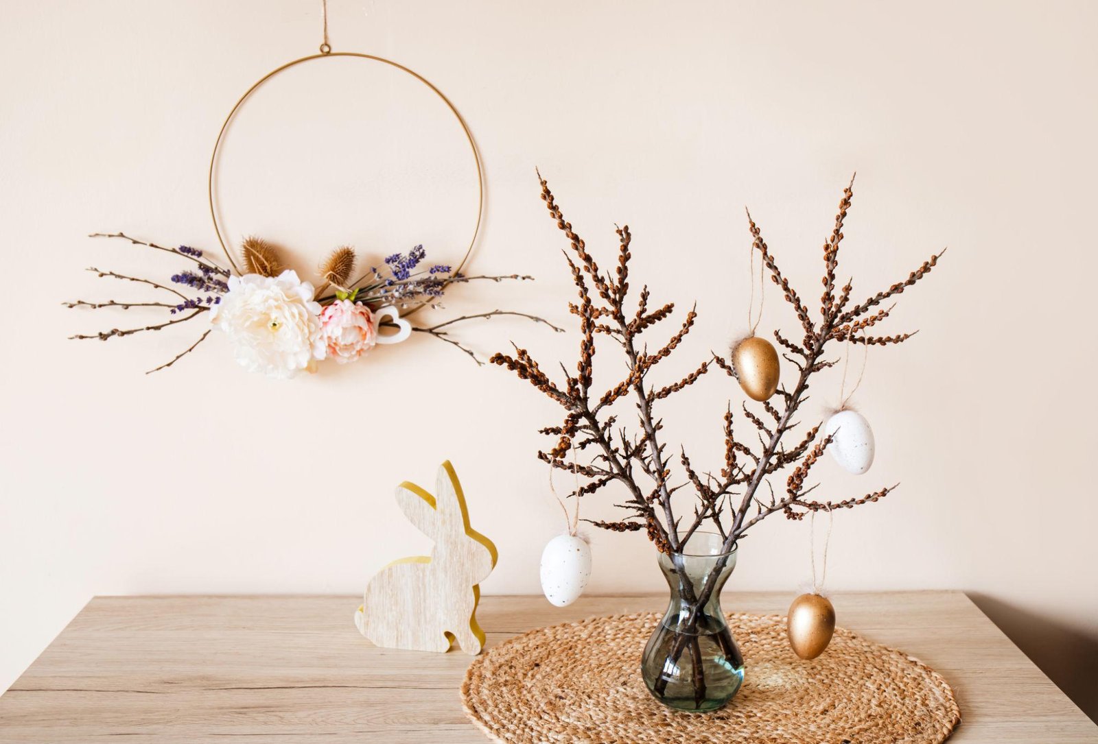 Minimalist Easter wreath with floral accents hanging on a wall, paired with a vase of branches decorated with hanging eggs and a wooden bunny on a rustic table.