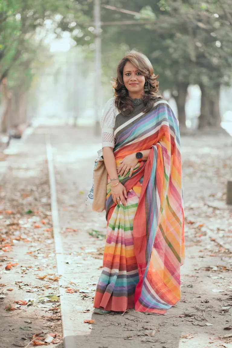 Woman wearing a colorful striped saree standing on a tree-lined path with soft sunlight, styled in a graceful and modern traditional look.