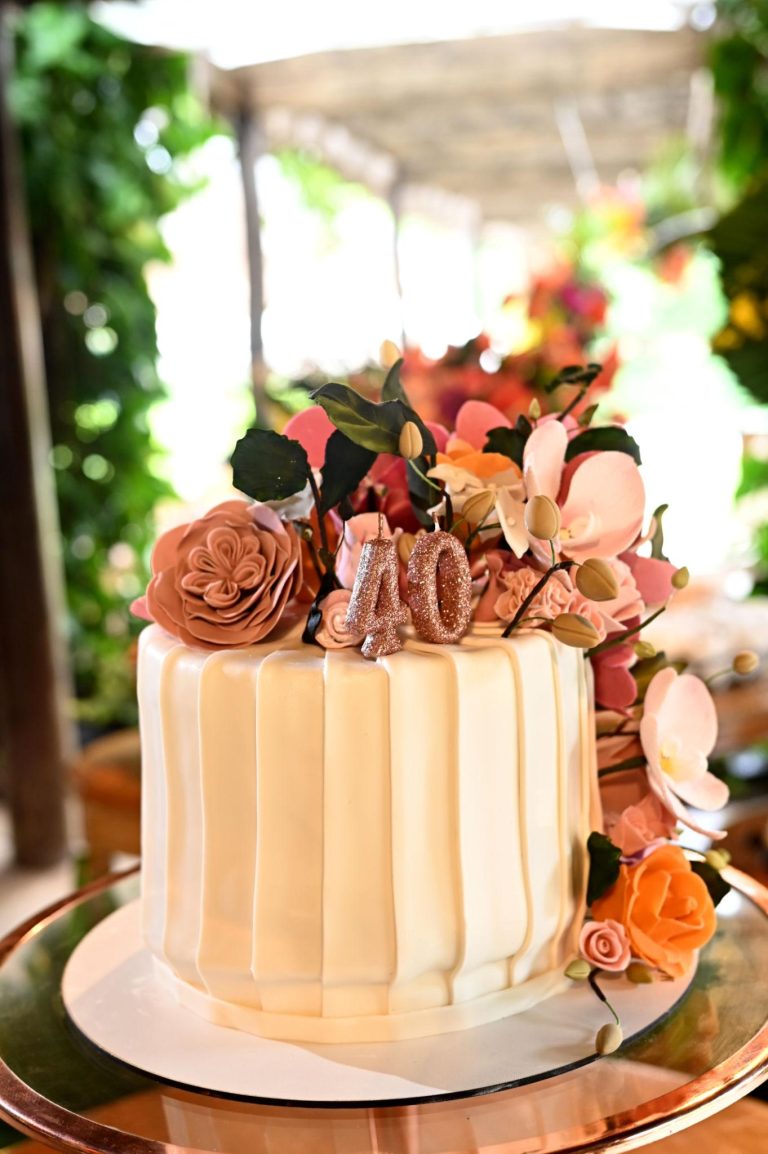 Elegant birthday cake with smooth white frosting, floral decorations in soft peach and pink tones, and a glittery number 40 topper set in a warm outdoor celebration setting.