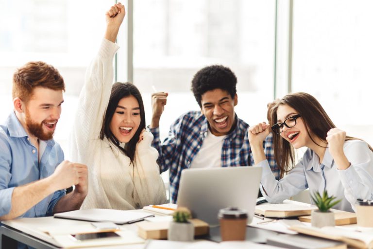 Group of happy students celebrating exam success together around a laptop, cheering with excitement in a bright classroom after receiving positive examination results.