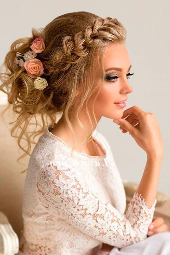 A beautiful blonde bride wears an elegant braided updo decorated with soft pink and cream roses. Loose curls frame her face while her lace wedding dress adds romantic charm to the sophisticated bridal hairstyle.