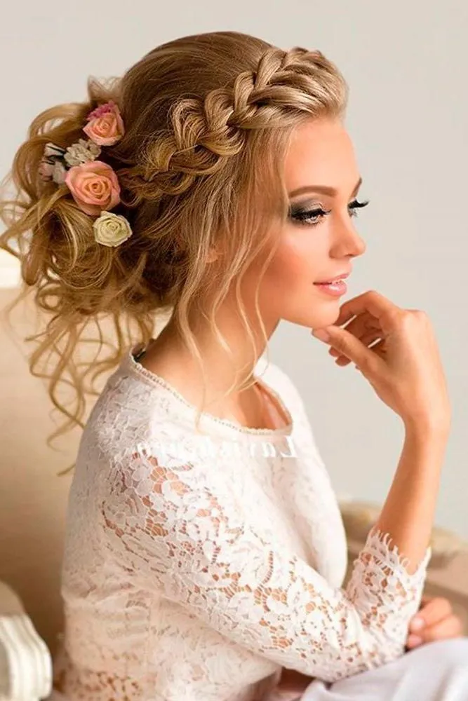 A beautiful blonde bride wears an elegant braided updo decorated with soft pink and cream roses. Loose curls frame her face while her lace wedding dress adds romantic charm to the sophisticated bridal hairstyle.