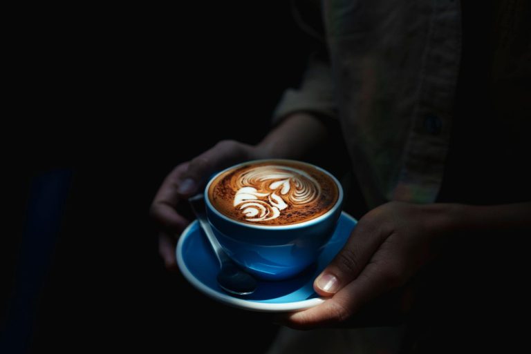 Close up of a warm latte with beautiful latte art served in a blue coffee cup, held by hands in a dark cozy setting, creating a calm and aesthetic coffee party atmosphere.