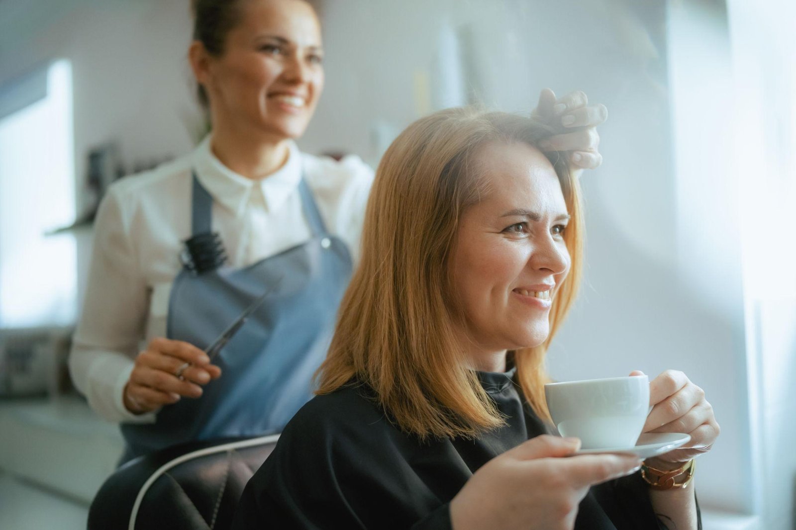 Woman enjoying a salon appointment while a hairstylist carefully examines and styles her shoulder-length hair. The client smiles while holding a cup of coffee, creating a relaxed and welcoming beauty salon atmosphere.