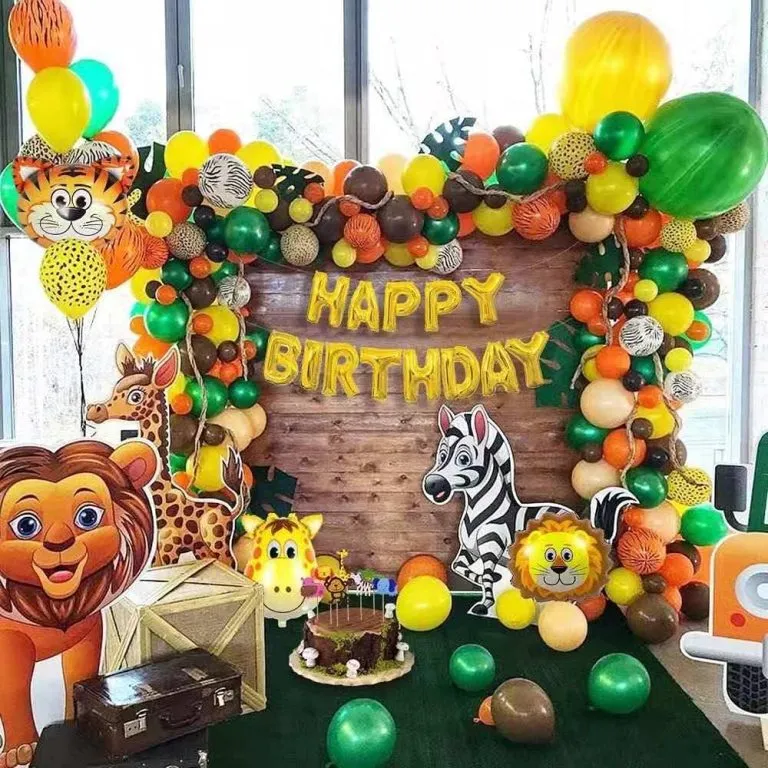 Colorful Safari Birthday Party backdrop featuring a jungle-themed balloon garland in green, yellow, orange, and brown tones, with cartoon lion, zebra, and giraffe cutouts, wooden crates, birthday cake display, and a rustic wooden background with Happy Birthday balloon lettering.