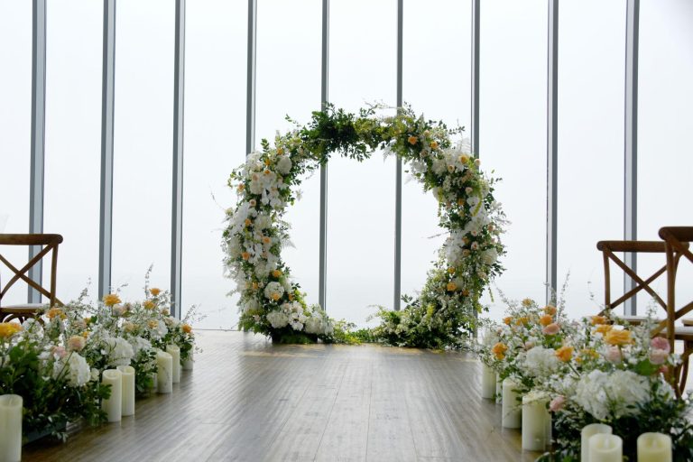 Elegant indoor wedding ceremony setup featuring a circular floral wedding arch adorned with white blooms, soft peach roses, and lush greenery. A wooden aisle is lined with flowers and white pillar candles, with large floor-to-ceiling windows creating a bright, romantic backdrop.