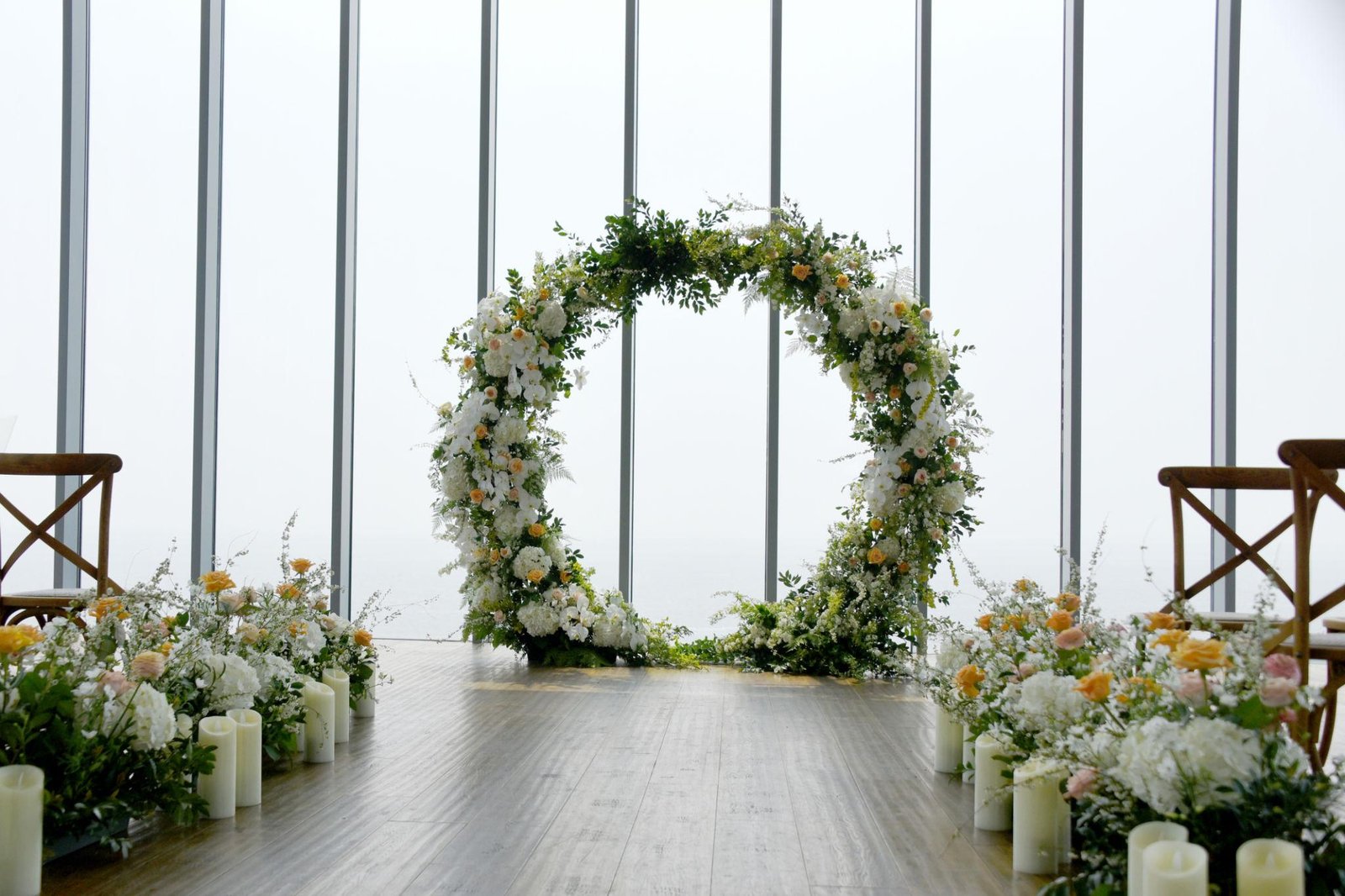 Elegant indoor wedding ceremony setup featuring a circular floral wedding arch adorned with white blooms, soft peach roses, and lush greenery. A wooden aisle is lined with flowers and white pillar candles, with large floor-to-ceiling windows creating a bright, romantic backdrop.