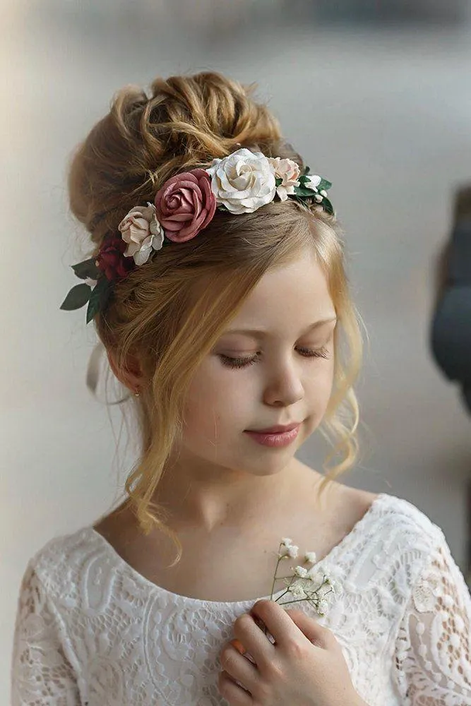 Young flower girl with a soft blonde updo hairstyle adorned with a floral crown of pink and white roses, wearing a white lace dress and holding delicate baby's breath flowers.