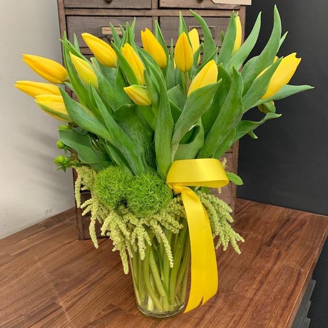 Elegant yellow tulip bouquet arranged in a clear glass vase with lush green foliage, decorative moss, and a yellow satin ribbon, displayed on a wooden table for modern home décor