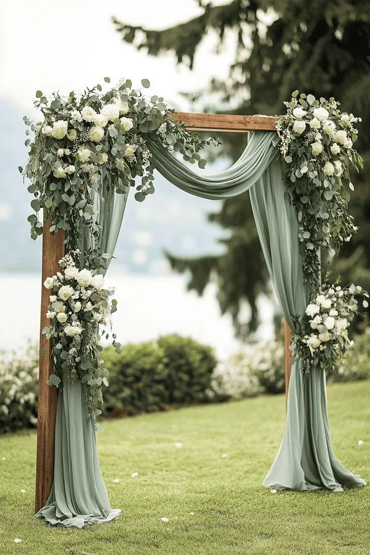Elegant sage green wedding backdrop featuring soft draped fabric, wooden arch frame, white roses, eucalyptus greenery, and romantic floral arrangements in an outdoor garden ceremony setting.