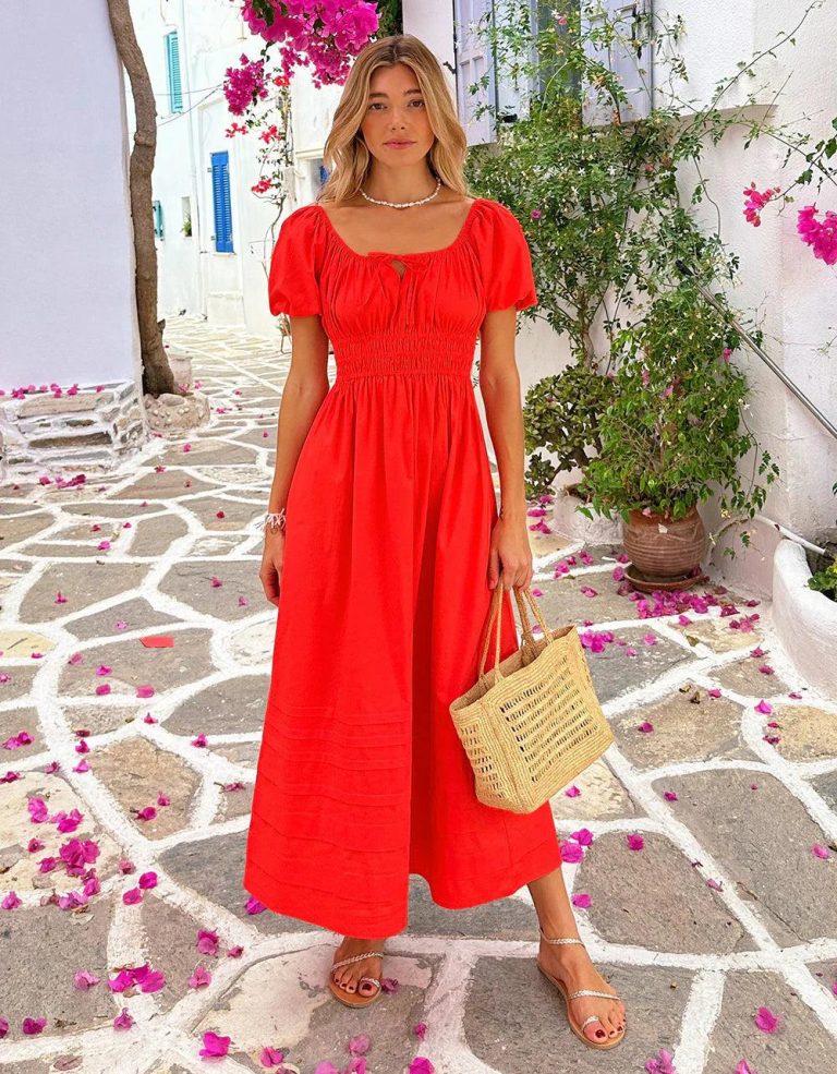 Tomato Girl Outfit Ideas featuring a woman wearing a white linen dress with a straw bag, espadrille sandals, gold jewelry, and a Mediterranean-inspired summer setting with olive trees and rustic architecture.