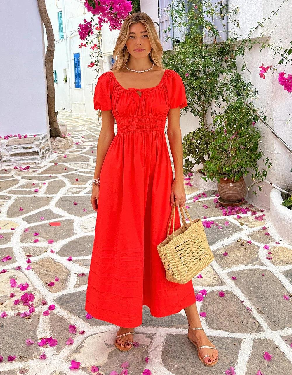 Tomato Girl Outfit Ideas featuring a woman wearing a white linen dress with a straw bag, espadrille sandals, gold jewelry, and a Mediterranean-inspired summer setting with olive trees and rustic architecture.