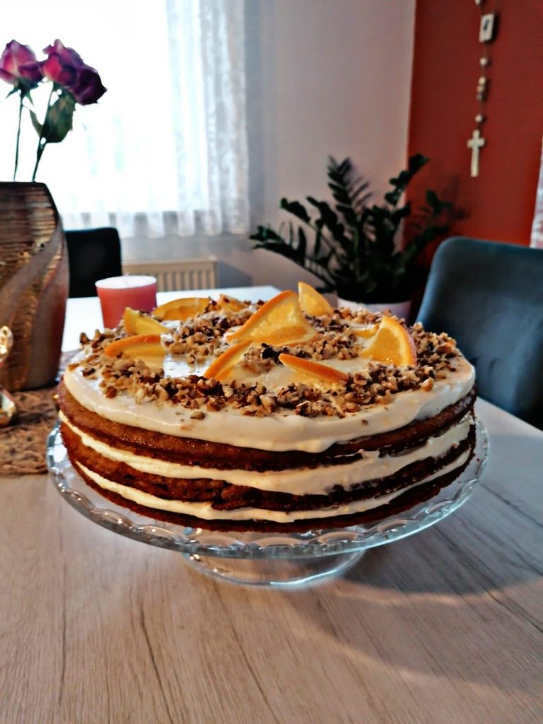 Moist carrot cake with orange cream cheese frosting, fresh orange slices, walnuts, and elegant decoration on a white cake stand. Perfect homemade dessert for birthdays, Easter, and afternoon tea.