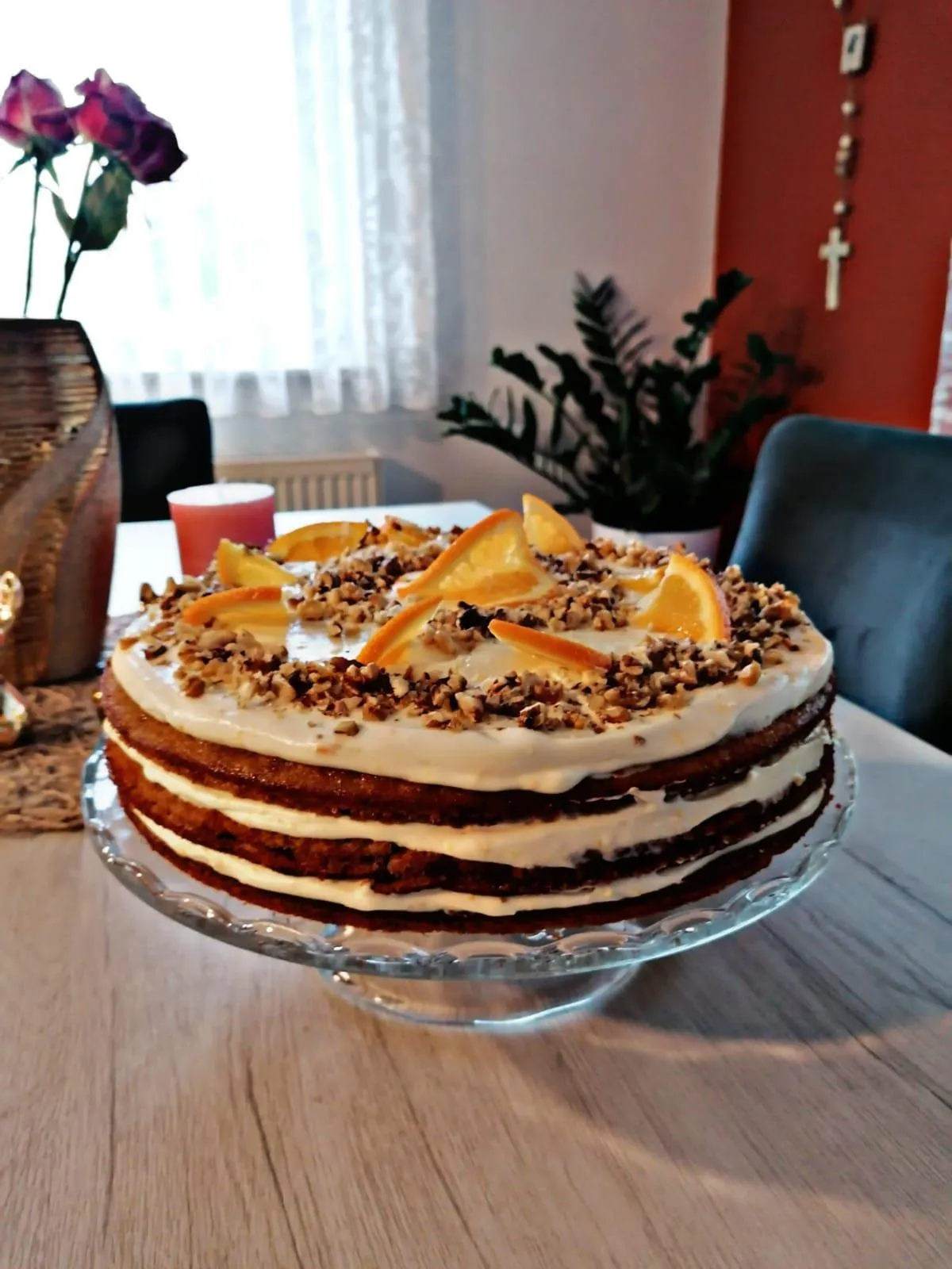 Moist carrot cake with orange cream cheese frosting, fresh orange slices, walnuts, and elegant decoration on a white cake stand. Perfect homemade dessert for birthdays, Easter, and afternoon tea.