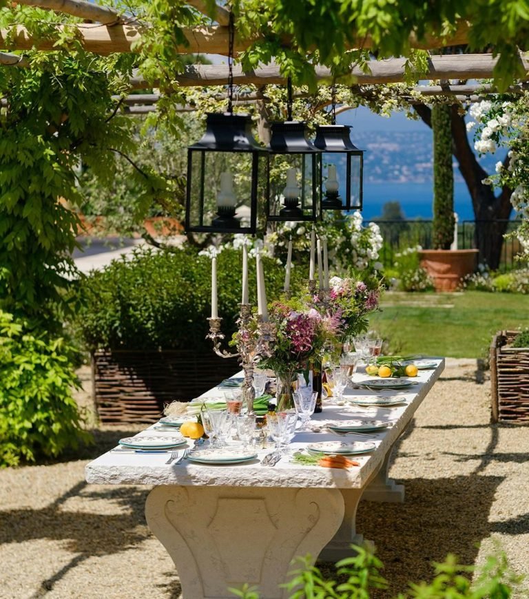 European Summer Party Ideas featuring a Mediterranean-style outdoor dining table with elegant floral centerpieces, hanging lanterns, candle holders, fresh lemons, and a luxury garden tablescape overlooking the coast.