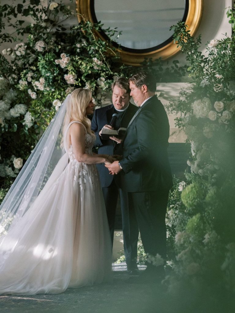 Intimate wedding ceremony featuring a bride and groom exchanging vows with an officiant, surrounded by lush white flowers and greenery in an elegant indoor setting.