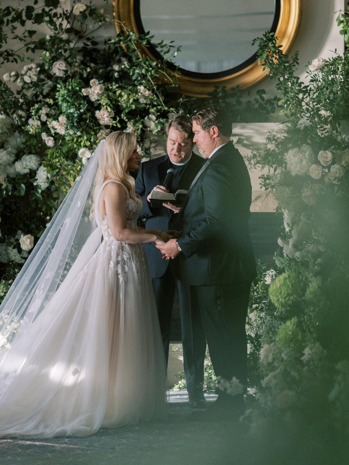Intimate wedding ceremony featuring a bride and groom exchanging vows with an officiant, surrounded by lush white flowers and greenery in an elegant indoor setting.