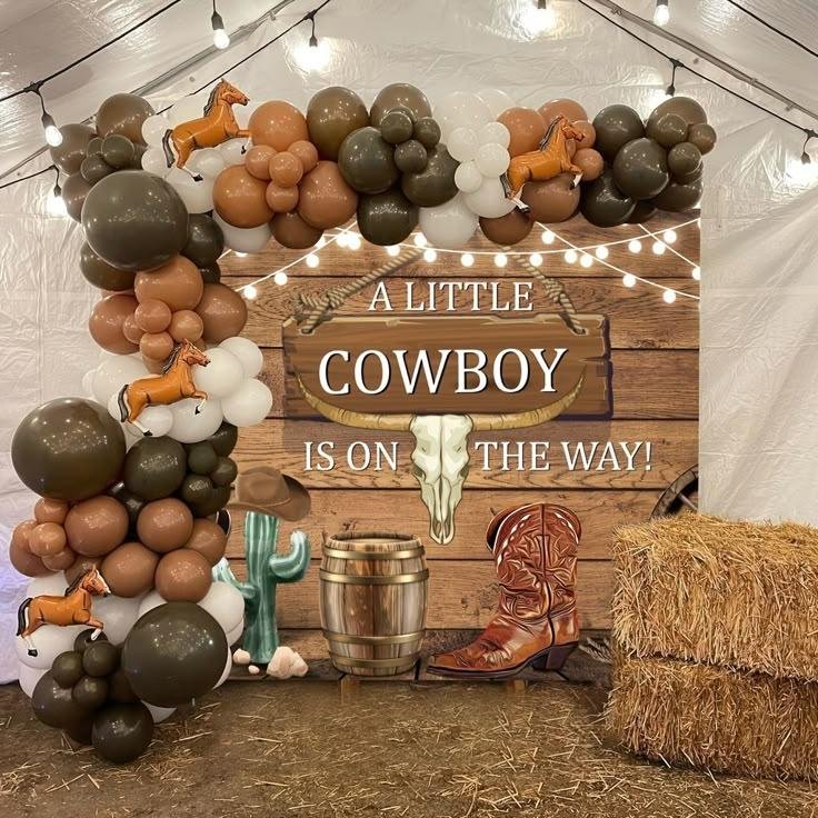 Rustic cowboy baby shower backdrop featuring a brown, white, and sage green balloon garland, wooden western sign, cowboy boots, cowboy hat, cactus, hay bales, barrel, and warm string lights.